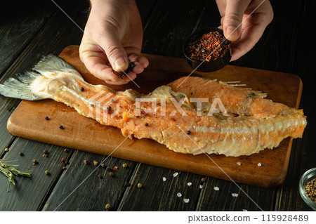 Cooking salmon fish on the kitchen table. A chef adds spices to a fish steak with his hands before baking in the oven 115928489