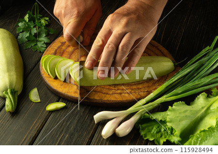 Close-up of a chef hands cutting zucchini into small pieces with a knife before frying or preparing a vegetarian dish. An idea for cooking zucchini with fresh vegetables and aromatic spices Close-up of a chef hands cutting zucchini into small pieces with a knife before frying or preparing a vegetarian dish. An idea for cooking zucchini with fresh vegetables and aromatic spices 115928494
