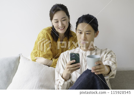 A middle-aged man sitting on a sofa in the living room looking at his smartphone and a middle-aged woman looking at the smartphone from behind A middle-aged man sitting on a sofa in the living room looking at his smartphone and a middle-aged woman looking at the smartphone from behind 115928545