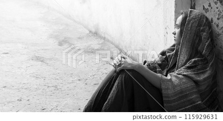 A girl in poor clothes sits on the ground 115929631