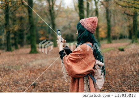 Woman wrapped in Shawl Poncho with backpack making photo by phone while enjoying autumn walk in forest. Capture the moment. Feeling harmony. Relaxing, personal fulfillment. Local travel lifestyle. 115930818