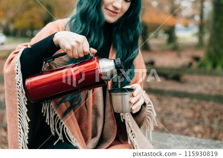 Woman wrapped in Shawl Poncho sits on picnic table pouring hot drink from thermos during autumn walk in forest with fall leaves. Feeling harmony, relax, personal fulfillment. Local travel lifestyle. Woman wrapped in Shawl Poncho sits on picnic table pouring hot drink from thermos during autumn walk in forest with fall leaves. Feeling harmony, relax, personal fulfillment. Local travel lifestyle. 115930819