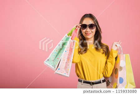 A cheerful Asian teen, elegantly holding shopping bags, smiles with excitement in this studio shot on a pink background. She's a portrait of joyful and pretty shopping. black friday 115933080