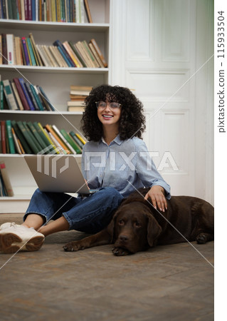 Cute woman working on laptop and stroking her dog lying next to her Cute woman working on laptop and stroking her dog lying next to her 115933504