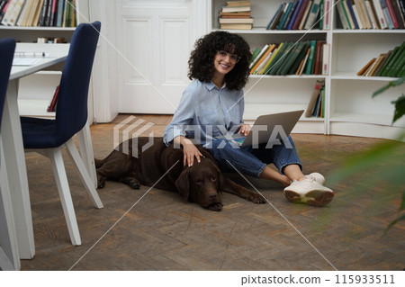 Curly-haired woman with laptop sitting on the floor, a dog next to her 115933511