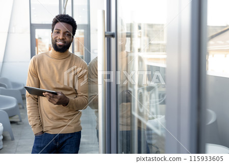 Positive african american guy with a tablet in hands standing outdoors 115933605