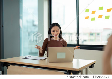 Smiling Call Center Agent Working on Laptop with Headset in Modern Office Environment Smiling Call Center Agent Working on Laptop with Headset in Modern Office Environment 115933677