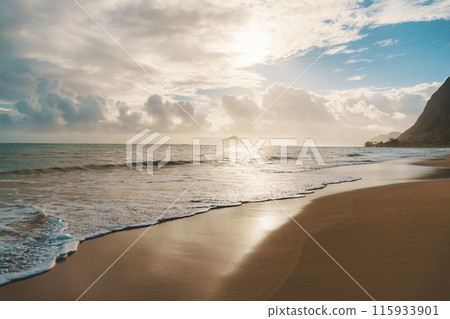 The sun sets, casting warm glow over the ocean on a cloudy day at the beach The sun sets, casting warm glow over the ocean on a cloudy day at the beach 115933901