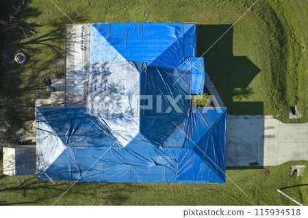 Aerial view of damaged in hurricane Ian house roof covered with blue protective tarp against rain water leaking until replacement of asphalt shingles Aerial view of damaged in hurricane Ian house roof covered with blue protective tarp against rain water leaking until replacement of asphalt shingles 115934518