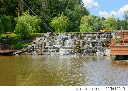 Artificial embankment with waterfall in the park on a sunny afternoon. Artificial embankment with waterfall in the park on a sunny afternoon. 115936643