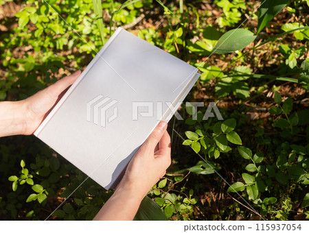 . Blank book hard back mock up in hand in nature, sunny day, green plants, grass. . Blank book hard back mock up in hand in nature, sunny day, green plants, grass. 115937054