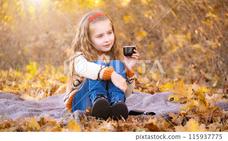 Nice little girl with cup sitting on picnic mat in autumn park Nice little girl with cup sitting on picnic mat in autumn park 115937775
