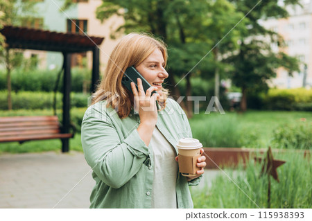 Happy cheerful young woman in fashion clothes talking on Phone with eco cup of tea. Mockup, Woman holding smartphone and coffee cup in city park. Urban, drinks and people concept. High quality photo 115938393