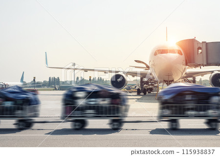 Luggage motion blurred trolley cart going fast delivering passenger baggage to modern plane on taxiway at airport on bright sunny day. Commercial aircraft on background at sunset or sunrise time 115938837