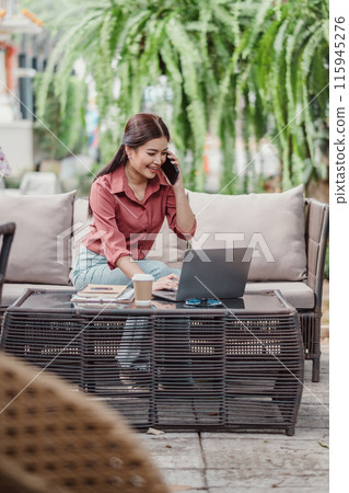 Young woman in casual business attire working on a laptop and talking on the phone while sitting outdoors in a garden setting. Young woman in casual business attire working on a laptop and talking on the phone while sitting outdoors in a garden setting. 115945276