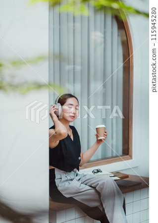 A young woman wearing headphones, holding a coffee cup, and relaxing by a window in a modern minimalist setting. 115945280