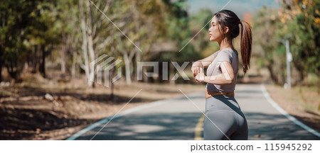 Young woman stretching on a scenic road surrounded by trees, preparing for a workout on a sunny day. 115945292