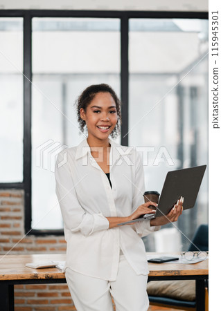 A young professional woman in a white outfit holding a laptop and a coffee cup, standing in a modern office with a brick wall and large windows. 115945301