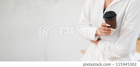 Close-up of a woman in a white shirt holding a coffee cup, showcasing her long manicured nails against a minimalist background. 115945302
