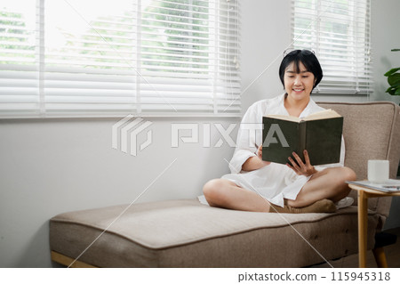 A woman enjoys a peaceful moment reading a book on a cozy sofa in a well-lit, modern living room with large windows and white blinds. A woman enjoys a peaceful moment reading a book on a cozy sofa in a well-lit, modern living room with large windows and white blinds. 115945318
