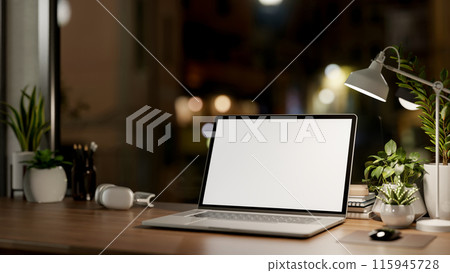 A laptop computer with a white-screen mockup on a wooden table in a dark room at night. A laptop computer with a white-screen mockup on a wooden table in a dark room at night. 115945728
