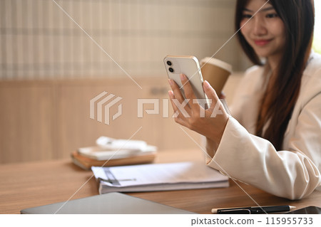 An attractive businesswoman drinking coffee and using smartphone at desk, checking social media or communicating online 115955733