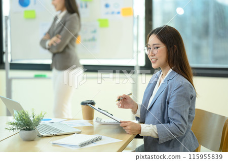 Portrait of smiling businesswoman working in modern office with colleagues in the background Portrait of smiling businesswoman working in modern office with colleagues in the background 115955839