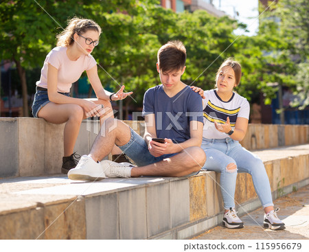 Teenage boy absorbed in phone, ignoring girls talking to him Teenage boy absorbed in phone, ignoring girls talking to him 115956679