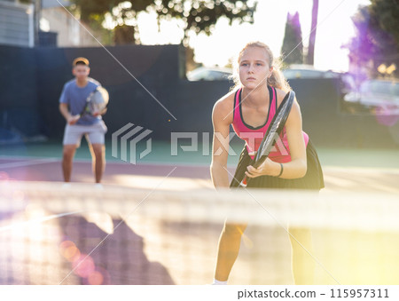 Young woman playing tennis doubles match with male partner on court at sunny day 115957311