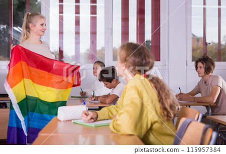 Progressive female teacher discussing with teenage students about LGBT social movements in classroom, holding rainbow flag 115957384