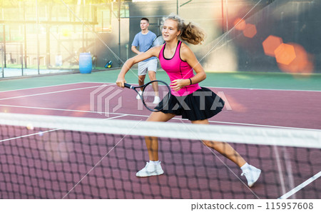 Positive female player hits ball close to net while playing tennis in rays of sun and glare 115957608