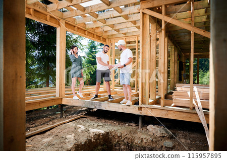 Man with detailed plan in hand showcases to couple the process of constructing wooden framed house near the forest. Men firmly grasp each other's hands in a gesture of mutual respect. Man with detailed plan in hand showcases to couple the process of constructing wooden framed house near the forest. Men firmly grasp each other's hands in a gesture of mutual respect. 115957895