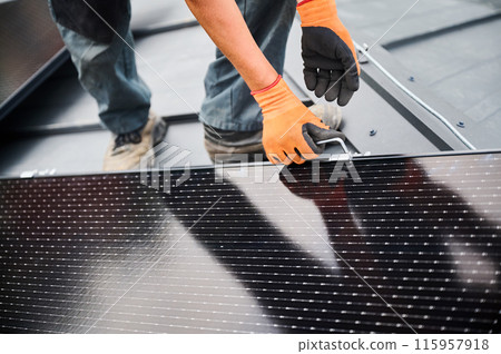 Worker building photovoltaic solar panel system on rooftop of house. Man engineer in gloves installing solar module with help of hex key outdoors. Alternative and renewable energy generation concept. 115957918