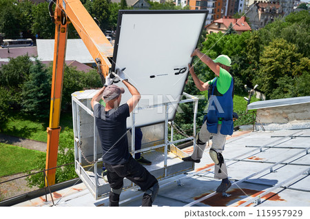 Workers lifting up photovoltaic solar panel on metal rooftop of house with assistance of crane lift. Men installers installing photovoltaic solar modules outdoors. Renewable energy generation concept. 115957929