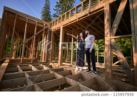 Man and woman examining their future wooden frame dwelling nestled near the forest. Youthful couple at construction site in early morning. Concept of contemporary ecological construction. 115957930