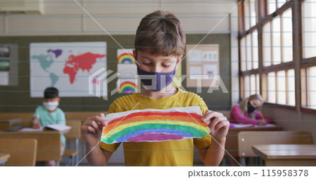 A Caucasian child in a yellow shirt and mask shows a rainbow drawing at school 115958378