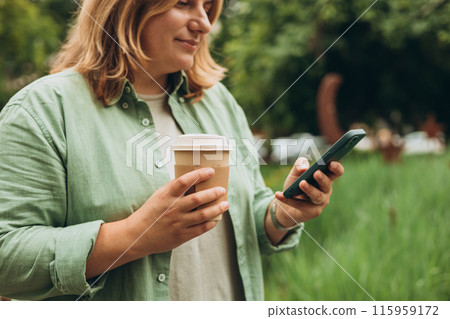 Beautiful smiling 30s female with coffee cup using smartphone and standing outdoors. Phone Communication. Happy cheerful young woman walking on city park, Urban lifestyle concept. 115959172