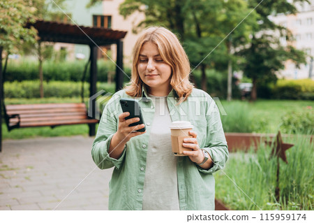 Beautiful smiling 30s female with coffee cup using smartphone and standing outdoors. Phone Communication. Happy cheerful young woman walking on city park, Urban lifestyle concept. 115959174