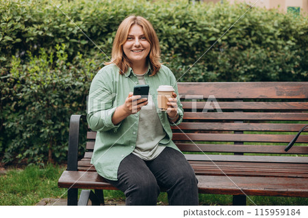 Beautiful smiling 30s female with coffee cup using smartphone and standing outdoors. Phone Communication. Happy cheerful young woman walking on city park, Urban lifestyle concept. Beautiful smiling 30s female with coffee cup using smartphone and standing outdoors. Phone Communication. Happy cheerful young woman walking on city park, Urban lifestyle concept. 115959184