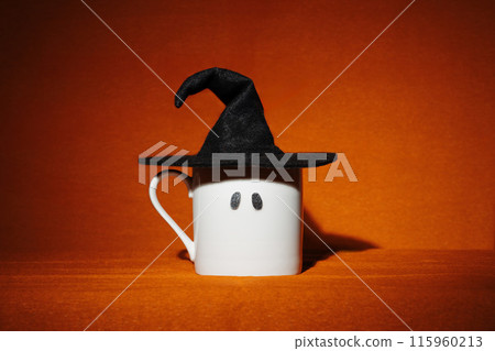 White mug with sparkly spooky ghost eyes and witch hat on orange background. Creative minimalist Halloween wallpaper 115960213