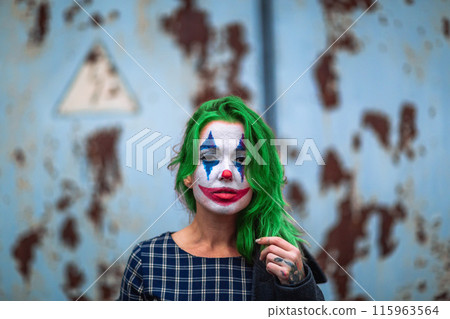 Portrait of a greenhaired girl in chekered dress with joker makeup on a rusty metal wall background. 115963564
