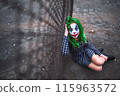 Greenhaired girl in chekered dress with joker makeup sitting near wire mesh fence on the ground. 115963572