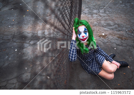 Greenhaired girl in chekered dress with joker makeup sitting near wire mesh fence on the ground. 115963572