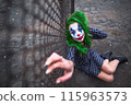 Greenhaired girl in chekered dress with joker makeup sitting near wire mesh fence on the ground. 115963573