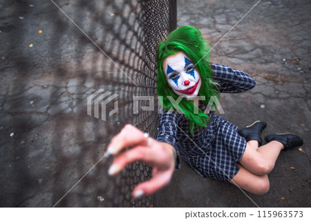 Greenhaired girl in chekered dress with joker makeup sitting near wire mesh fence on the ground. 115963573