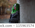 Portrait of a greenhaired girl in chekered dress with joker makeup on a brick wall background. 115963574