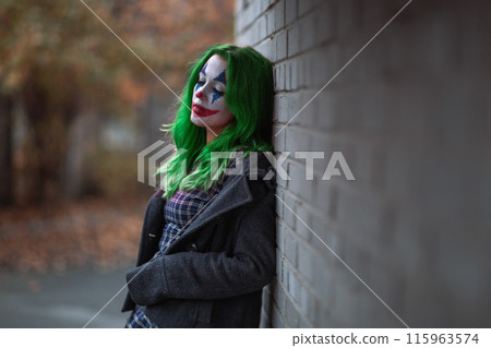 Portrait of a greenhaired girl in chekered dress with joker makeup on a brick wall background. 115963574