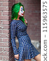 Portrait of a greenhaired girl in chekered dress with joker makeup on a brick wall background. 115963575