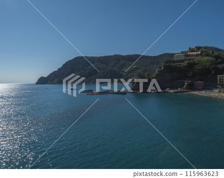 Picturesque distant view of Monterosso al Mare with beach, ships and boats seen from Azure Trail to Vernazza with green cliffs, rock, clear blue sky and sea. National park Cinque Terre, Liguria, Italy Picturesque distant view of Monterosso al Mare with beach, ships and boats seen from Azure Trail to Vernazza with green cliffs, rock, clear blue sky and sea. National park Cinque Terre, Liguria, Italy 115963623