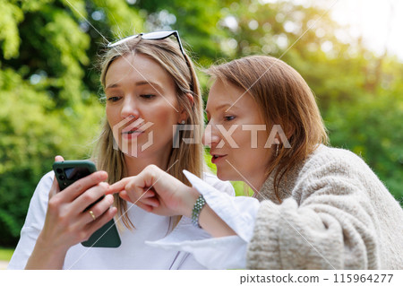 Two young adult happy smiling woman friends portrait using mobile phone smartphone outdoors. Female person people blogger girlfriend watching video streaming chating social media cell at city park Two young adult happy smiling woman friends portrait using mobile phone smartphone outdoors. Female person people blogger girlfriend watching video streaming chating social media cell at city park 115964277